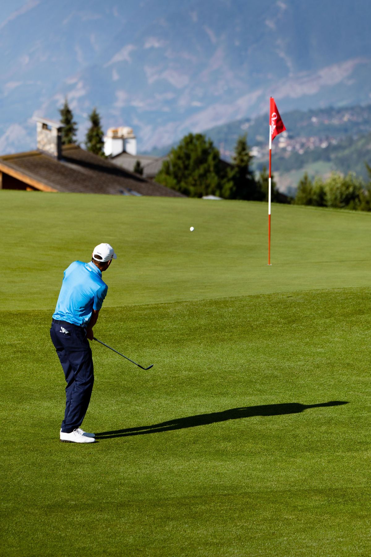 A golfer chipping onto an alpine green with mountains beyond